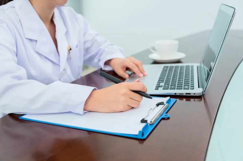 Doctor Working on Laptop and Clipboard at Desk