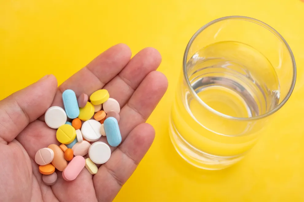 Hand holding assorted pills next to glass of water on yellow background