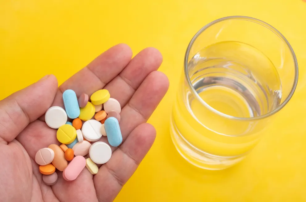 Hand holding pills next to glass of water over yellow background