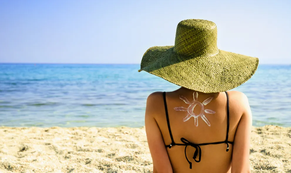 Woman sitting on beach with wide brimmed hat and sun cream on back